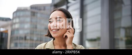 Prise de vue verticale d'une femme asiatique souriante dans un casque sans fil, aime écouter de la musique dans un casque, tient le téléphone portable Banque D'Images