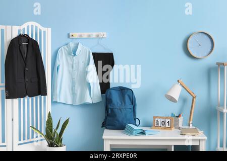 Intérieur du lieu de travail avec bureau, écran pliant, sac à dos, horloge et uniforme scolaire élégant accroché sur le mur bleu Banque D'Images