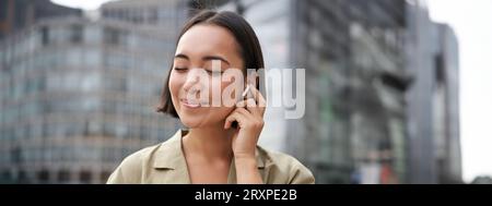 Prise de vue verticale d'une femme asiatique souriante dans un casque sans fil, aime écouter de la musique dans un casque, tient le téléphone portable Banque D'Images