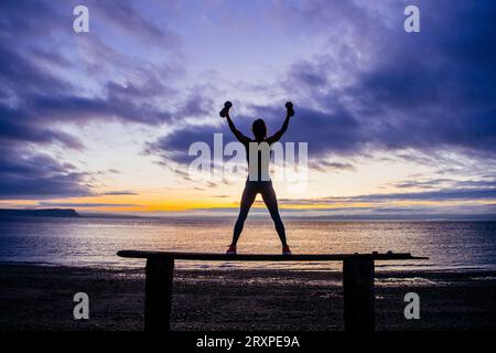 Silhouette de femme s'exerçant avec des haltères sur le dessus du banc côtier au crépuscule nuageux Banque D'Images