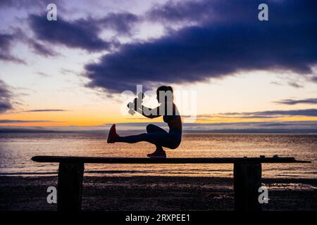 Silhouette de femme s'exerçant avec des haltères sur le dessus du banc côtier au crépuscule nuageux Banque D'Images