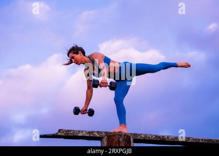 Silhouette de femme s'exerçant avec des haltères sur le dessus du banc côtier au crépuscule nuageux Banque D'Images
