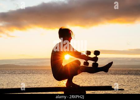 Silhouette de femme s'exerçant avec des haltères sur le dessus du banc côtier au crépuscule nuageux Banque D'Images