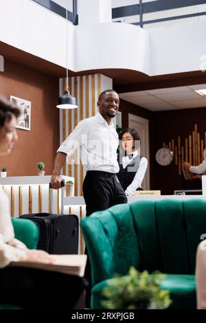 Invité masculin assis à la réception pour remplir les dossiers d'inscription, personne souriant et attendant l'hébergement à l'hôtel. Jeune touriste debout près de la réception dans le hall, complexe moderne. Banque D'Images
