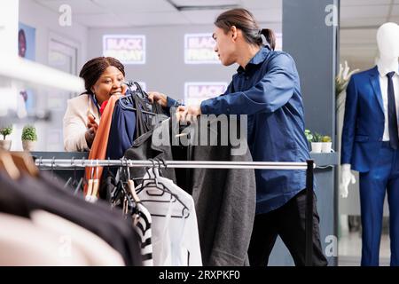 Amicale souriante femme afro-américaine travailleuse au détail aidant le client masculin à choisir des vêtements le Black Friday. Caissière féminine agréable servant l'acheteur dans le magasin de vêtements pendant la saison des soldes Banque D'Images