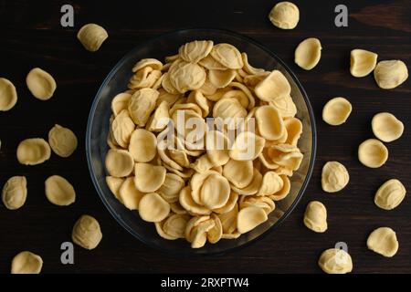Pâtes jaunes séchées à l'orecchiette dans un bol en verre : un bol de pâtes orecchiette non cuites sur une table en bois vue du dessus Banque D'Images