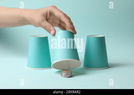 Femme montrant des pièces de monnaie sous la tasse sur fond bleu clair, closeup. Jeu Thimblerig Banque D'Images