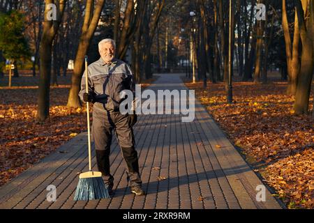 Travailleur masculin mature posant à la caméra avec balai dans le parc. Vue de face de l'homme barbu heureux en âge, portant l'uniforme debout avec balai et souriant, avec allée de parc sur fond. Concept de lieu de travail. Banque D'Images