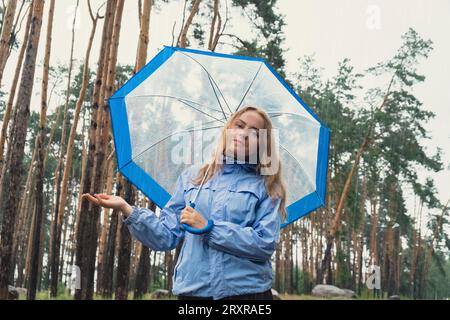 Fille blonde vérifiant le temps sous un parapluie transparent à l'extérieur dans le parc. Heure d'automne pluvieux mauvaises conditions de prévision saison. Touriste avec parapluie voyage dans de nouveaux endroits sous la pluie. Paysage Banque D'Images