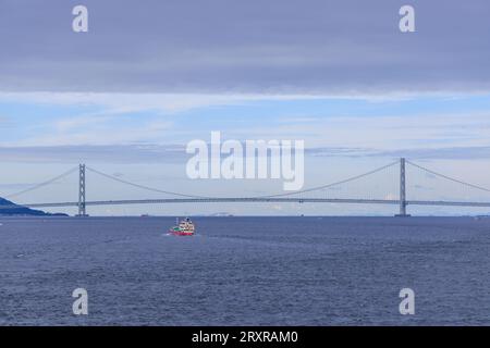 Petit navire navigue dans les eaux bleues calmes par pont suspendu par jour nuageux Banque D'Images