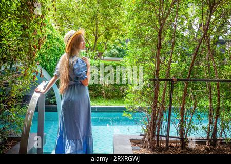 Femme en chapeau de paille et robe apprécie le matin par une piscine bleue Banque D'Images