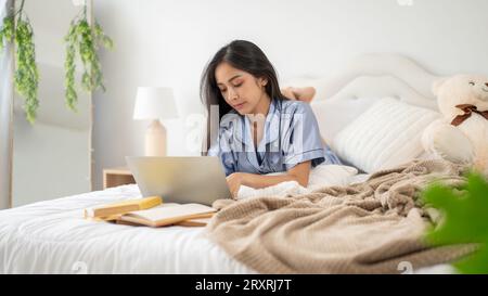 Une belle femme asiatique en pyjama se concentre sur son travail sur son ordinateur portable tout en étant couchée sur son lit dans sa chambre. Banque D'Images