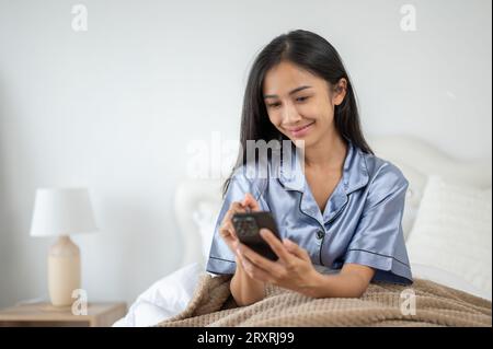 Une belle femme asiatique utilise son smartphone, vérifie les messages et répond par son smartphone tout en étant assise sur son lit après m'être réveillée Banque D'Images
