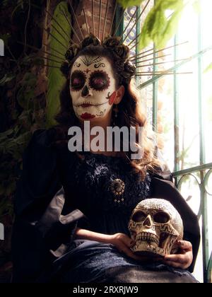 Femme mexicaine habillée en catrina pour le jour des morts, assise à côté d'une porte avec un crâne dans les mains. Banque D'Images