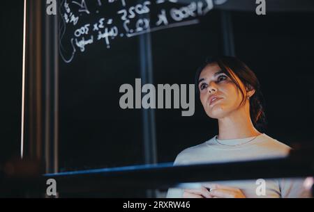 Pensive businesswoman contemple des idées derrière un mur de verre dans un bureau tard dans la nuit. Cet analyste de données créatif et entrepreneur travaille avec diligence, str Banque D'Images