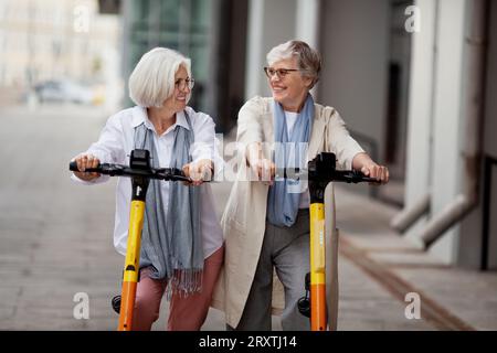 Portrait Happy Smiling Senior femmes avec les cheveux gris appréciant monter ensemble sur des scooters électriques à l'extérieur. Deux amies à la retraite se regardent Banque D'Images