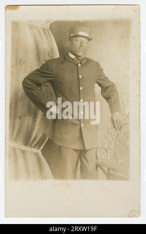 Carte postale photographique en noir et blanc qui n'a jamais été tamponnée et postée mais qui a écrit au crayon au dos le nom 'T.R. Joseph'. L'image sur la carte postale est de Pullman porter, T.R. Joseph debout dans un studio de photographie avec un fond peint d'un rideau de théâtre derrière lui. Il est vêtu d'un uniforme Pullman porter complet, avec chapeau Pullman porter, la main gauche sur la hanche et la main droite reposant sur une chaise en métal ornée. Banque D'Images