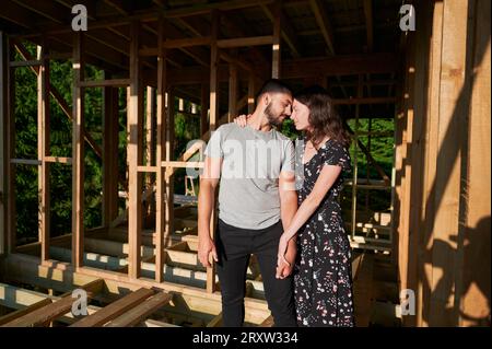 Homme et femme inspectant leur future habitation de cadre en bois nichée près de la forêt. Jeune couple sur le chantier en début de matinée. Concept de construction écologique contemporaine. Banque D'Images