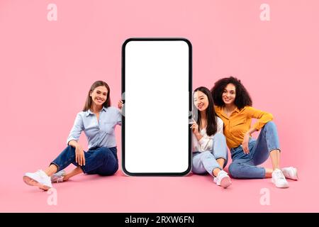Heureux trois amies féminines multiraciales assis près du téléphone portable géant avec écran blanc blanc, maquette, espace libre Banque D'Images
