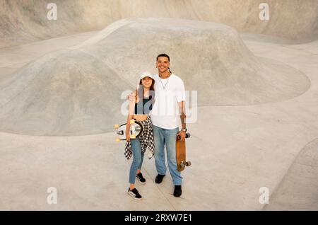 Jeune femme et gars posant avec des planches à roulettes et embrassant, se relaxant après des exercices sur rampe, pleine longueur, espace de copie Banque D'Images