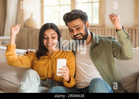 Joyeux couple indien utilisant smartphone et secouant les poings, lisant de bonnes nouvelles, célébrant la grande chance à la maison Banque D'Images
