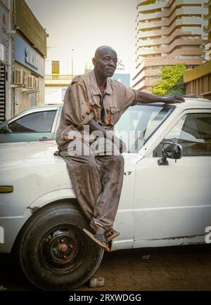 Une photo d'un Africain travaillant comme mécanicien assis sur sa voiture et portant des vêtements de travail Banque D'Images