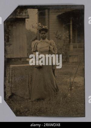 Tintype représentant une femme qui se tient dehors devant une maison et un hangar. Il y a une table drapée ou similaire derrière elle sur laquelle elle se penche légèrement en arrière. La femme porte une robe à manches bouffantes. Le haut semble clair et sa jupe sombre. La femme porte un chapeau et tient un petit sac dans sa main droite. La main gauche de la femme pend à son côté. Banque D'Images