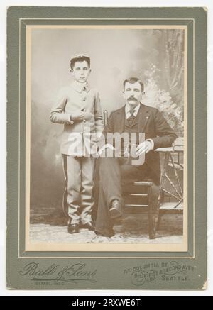 Photographie en noir et blanc d'un homme et d'un jeune garçon photographiés au studio ball & Sons de Seattle, WA. La photo est montée sur une carte d'armoire de couleur foncée estampillée avec le logo ball & Sons et l'emplacement du magasin sur le bas de la face avant. L'homme sur la photo est assis à gauche du garçon et porte un costume sombre. Son genou droit est croisé sur sa gauche et il tient un petit livre contre sa jambe gauche avec son coude reposant contre une table. Le garçon se tient à côté de lui et porte ce qui semble être un uniforme gris de style militaire et une casquette. La main droite est placée dans la veste reposant entre deux Banque D'Images