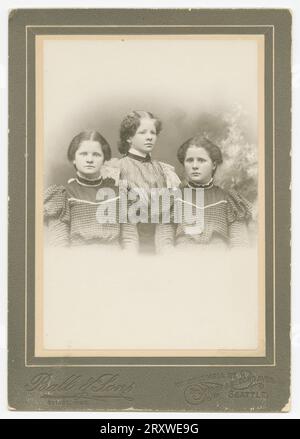 Une photographie en noir et blanc de trois filles photographiées au studio ball & Sons de Seattle, WA. La photo est montée sur une carte d'armoire de couleur foncée estampillée avec le logo ball & Sons et l'emplacement du magasin sur le bas de la face avant. Le trio est photographié de la taille vers le haut, avec deux filles assises et la troisième debout entre et juste derrière les deux autres. Les deux filles assises portent des robes de couleur foncée identiques et la troisième fille porte une robe plus légère avec des décorations en dentelle sur la manche. Les filles ont toutes les cheveux foncés qui ont été tirés vers l'arrière. Il y a une inscription au crayon o Banque D'Images