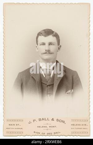 Une photographie en noir et blanc d'un homme photographié au studio J. P. ball & son à Helena, MT. La photo est montée sur une carte d'armoire de couleur claire avec des bords décoratifs et le nom et l'emplacement du studio photo imprimé sur le devant le long du fond. Le portrait est vignetté et montre un homme en veste sombre, gilet et cravate regardant directement l'appareil photo. Il a les cheveux bruns courts et une moustache. Banque D'Images