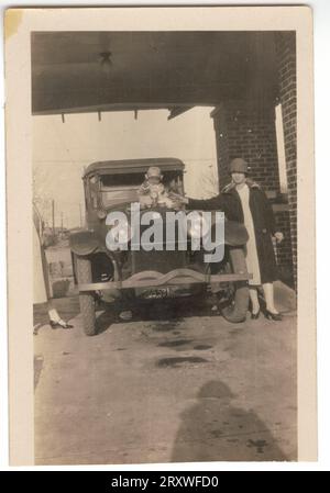 Une photographie en noir et blanc de deux (2) femmes debout à côté d'une voiture garée, avec un bébé assis sur le capot de la voiture. Au centre de la photo, le bébé Samuel Jackson Jr. Est posé assis sur le capot de la voiture. Il a le bras droit levé et il semble tendre la main vers l'ornement du capot de la voiture. Sur le côté gauche de la voiture, Mme Jackson se tient face à la caméra, la main droite tendue pour empêcher le bébé de tomber. Elle porte une robe de couleur claire, un pardessus de couleur foncée, des bas, des talons et un chapeau. Il y a une femme non identifiée à gauche, qui est presque completel Banque D'Images