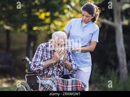 Homme âgé en fauteuil roulant éternuant dans la serviette pendant que la jeune infirmière s'occupe de lui dans le parc Banque D'Images