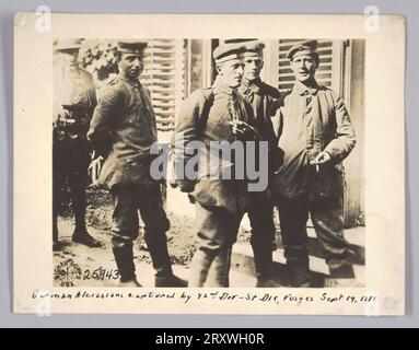Cette photographie représente quatre soldats capturés debout devant un bâtiment avec des volets. À l'extrême gauche de la photo, un soldat portant un casque et une veste ouverte se tient à une courte distance des soldats capturés. Les soldats portent des bérets et des vestes fermées. Un des soldats à gauche regarde directement la caméra. Deux soldats tiennent des cigarettes dans leurs mains et le soldat debout derrière eux regarde la caméra. Le logo signal corps USA se trouve dans le coin inférieur gauche de la photo. Le numéro [25943] est écrit à côté du logo. Le mot Banque D'Images