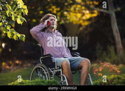 Jeune homme malheureux en fauteuil roulant parlant sur le téléphone portable dans le parc Banque D'Images