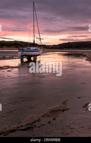 Yachts échoués attendant la marée assis sur le sable dans un estuaire au coucher du soleil avec la lumière reflétée dans le sable humide des nuages colorés du coucher du soleil Banque D'Images