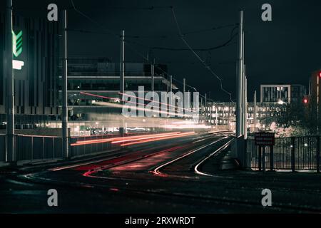 Mouvement flou des tramways circulant sur les rails à Nottingham City la nuit avec des sentiers de tramway légers et des bâtiments urbains éclairés en arrière-plan Banque D'Images