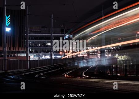 Mouvement flou des tramways circulant sur les rails à Nottingham City la nuit avec des sentiers de tramway légers et des bâtiments urbains éclairés en arrière-plan Banque D'Images