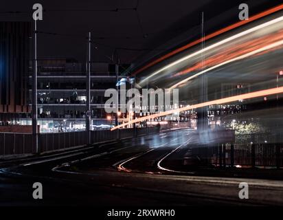 Mouvement flou des tramways circulant sur les rails à Nottingham City la nuit avec des sentiers de tramway légers et des bâtiments urbains éclairés en arrière-plan Banque D'Images