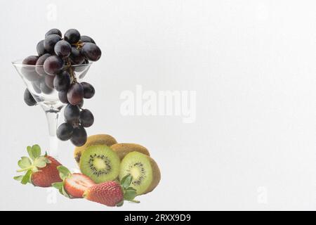 Raisins noirs succulents dans un verre à cocktail, accompagnés de fraises succulentes et de kiwi, présentés sur un fond blanc immaculé, laissant de la place Banque D'Images