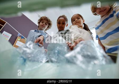 Groupe diversifié d'enfants mettant des bouteilles en plastique dans le bac de recyclage POV Banque D'Images