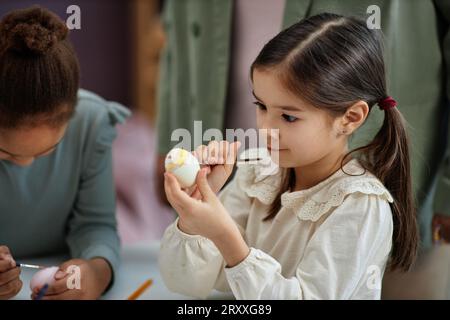 Portrait vue de côté de mignonne petite fille peignant soigneusement oeuf de Pâques dans la maternelle Banque D'Images