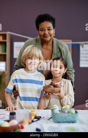 Portrait vertical de Kind Black Woman comme enseignante féminine posant avec des petits enfants mignons dans la maternelle tout en regardant la caméra Banque D'Images