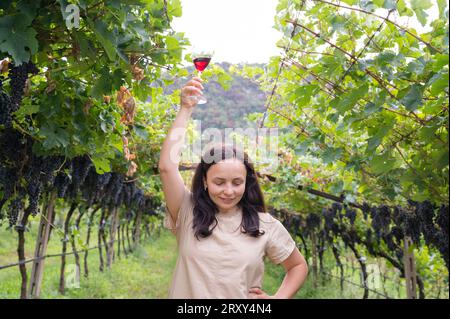 Belle femme rêvant de déguster du vin rouge en appréciant un séjour d'été dans les vignobles par belle journée ensoleillée. femme buvant du vin rouge au vignoble. saison de récolte. Photo de haute qualité Banque D'Images