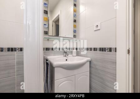 Petite salle de bains avec meubles en bois blanc, lavabo en porcelaine, armoire de miroir, miroir avec cadre coloré, cabine de douche blanche avec écrans et robinets chromés Banque D'Images