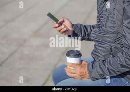 Détail des mains d'un homme latin tenant une tasse jetable avec du café et un téléphone portable. Banque D'Images