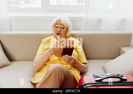 Une femme âgée heureuse avec passeport et billet de voyage a emballé une valise rouge, des vacances et des soins de santé. Une vieille femme souriante s'assoit volontiers sur le canapé avant Banque D'Images