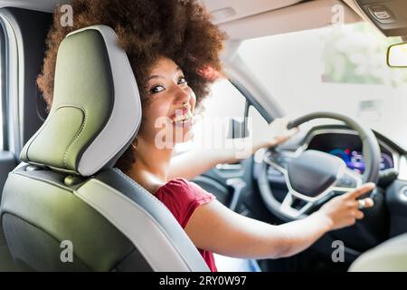 Heureuse femme afro-américaine avec coiffure afro conduisant l'automobile moderne tout en regardant loin avec sourire toothy Banque D'Images