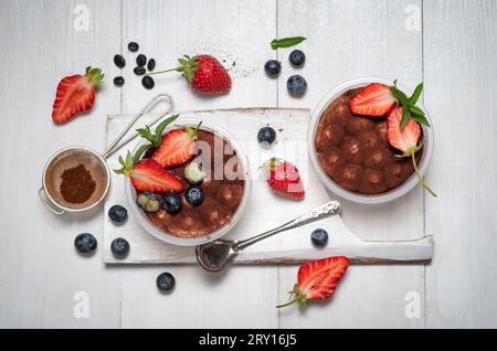 Dessert sucré italien traditionnel de gâteau tiramisu en verre sur une table en bois blanc. Avec de la poudre de cacao et un ajout rafraîchissant de fraise et de bleuet Banque D'Images