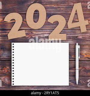 Numéros de papier cartonné 2024 Happy New Year Sign Near White Spiral Paper Cover Notebook avec stylo sur fond de table en bois. Rendu 3D. Banque D'Images