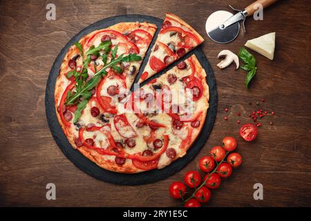 Pizza avec saucisses fumées, fromage, champignons, tomates cerises, poivrons et légumes verts sur une pierre et une planche en bois brun, vue de dessus. Banque D'Images